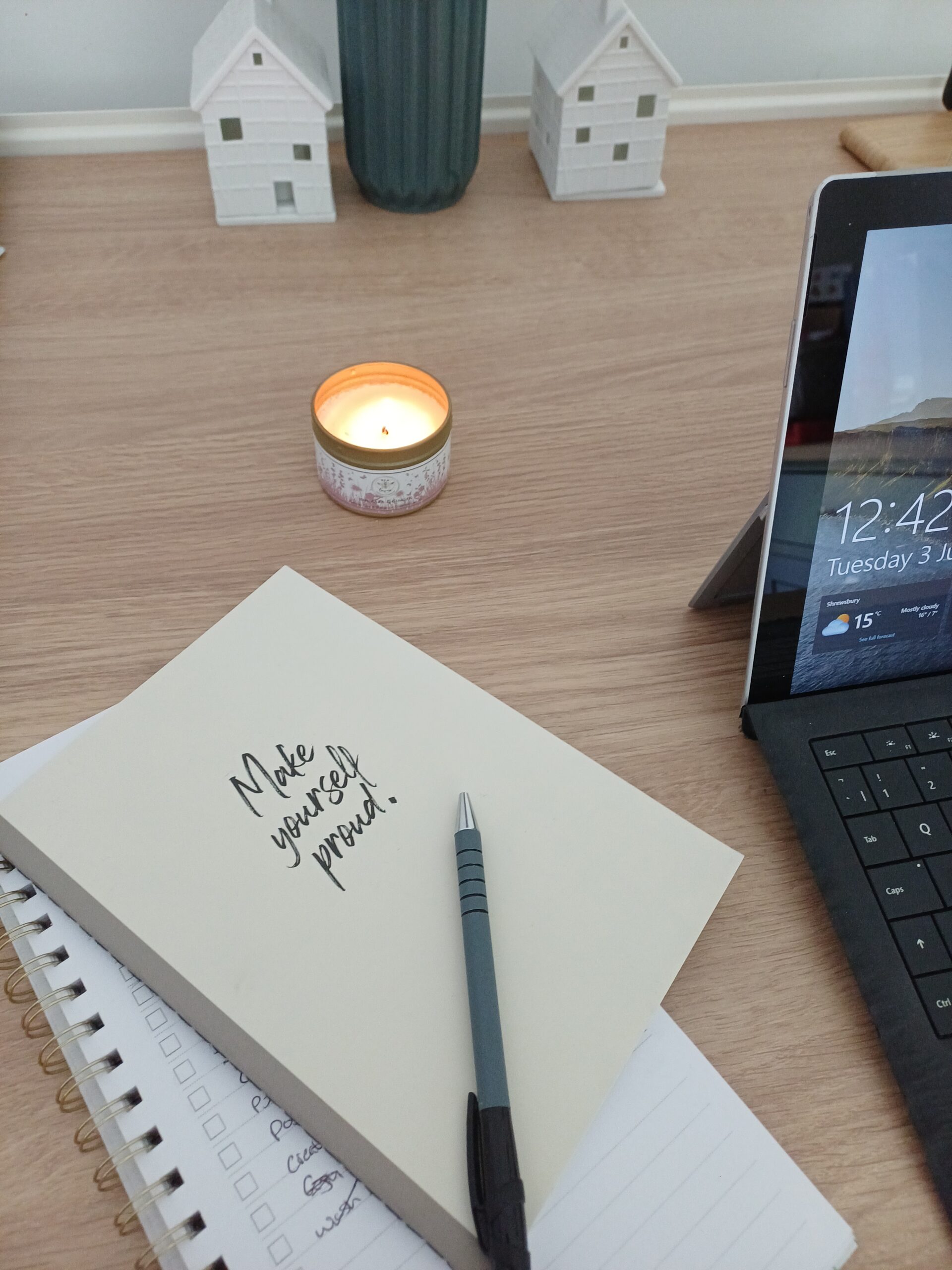 A candle is lit in the middle of a kitchen table. Note books, a laptop and decor pieces can be seen around.